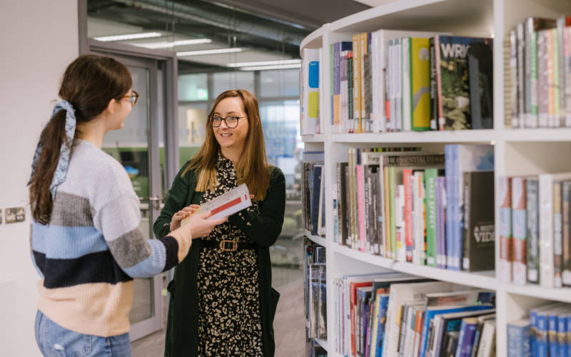 Two students talking beside a bookcase